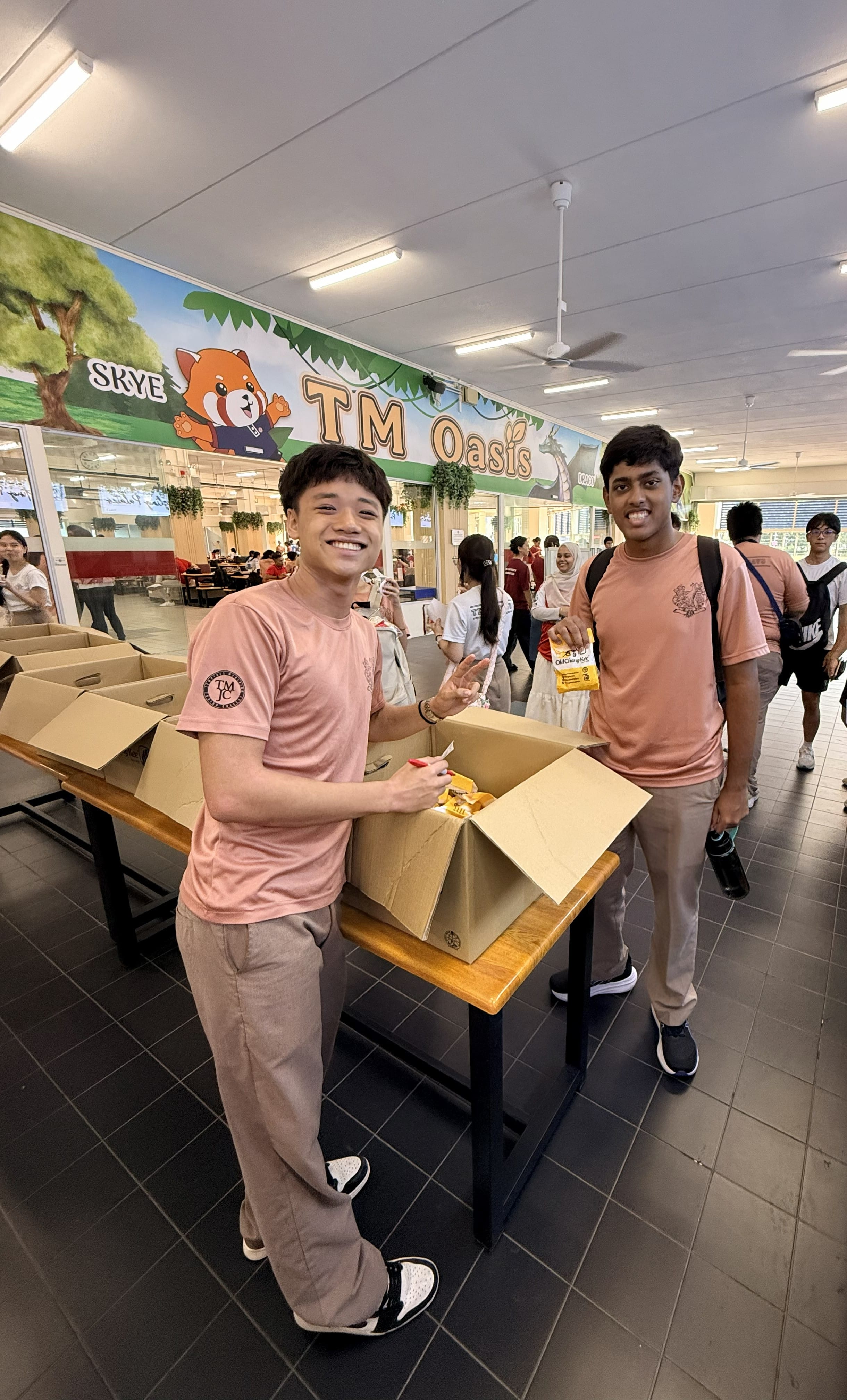 Two smiling students in matching pink T-shirts distribute snacks from open cardboard boxes at a school canteen, with a colourful "SKYE TM Oasis" mural featuring a cartoon red panda visible behind them.